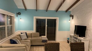 A living room with a beige sofa, a television on a white stand, and a sliding glass door leading to a balcony. - Beautiful House on the beach. Come and enjoy your paradise vacation! (George Town)