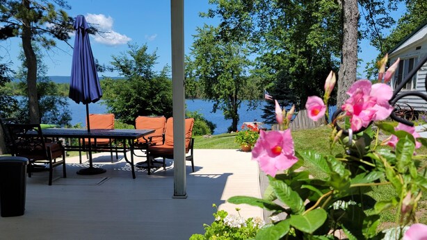 Un patio cubierto con mobiliario exterior, vista a un lago y macetas con flores rosadas.