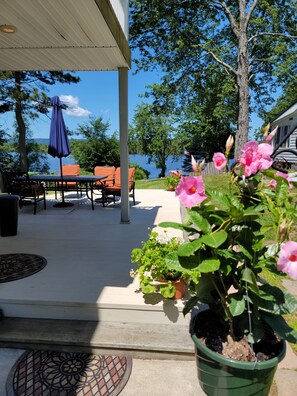 Un patio cubierto con mobiliario exterior, vista a un lago y macetas con flores rosadas.
