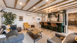 Una sala de estar moderna con techo de madera, una zona de comedor con taburetes de bar y una cocina con refrigerador de acero inoxidable.