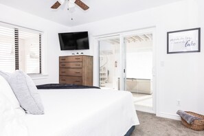 A bedroom with a bed, a TV, a wooden dresser, a sliding glass door to a balcony, and a framed quote on the wall.