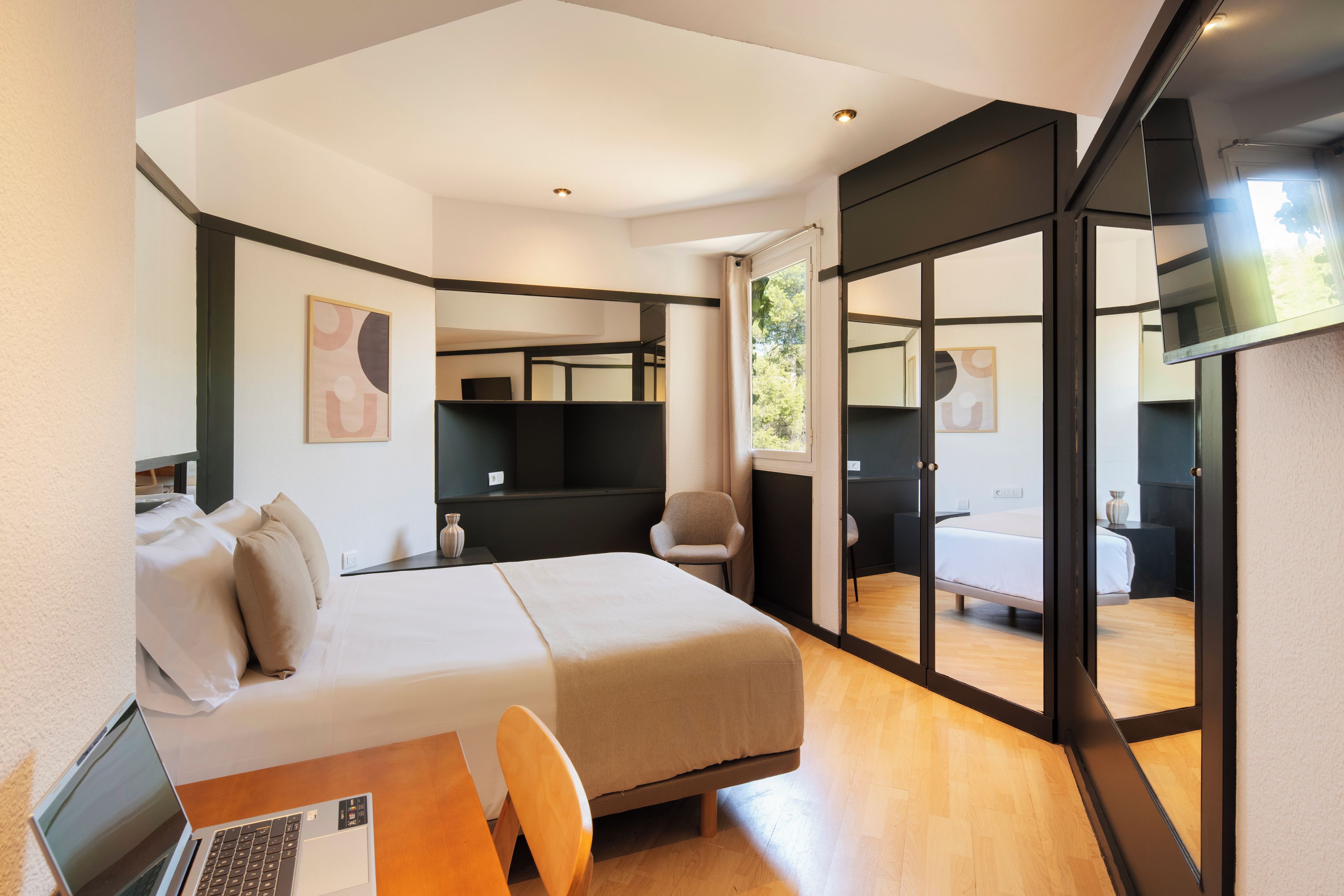A modern hotel room with a bed, a desk with a laptop, a chair, and a large mirror.