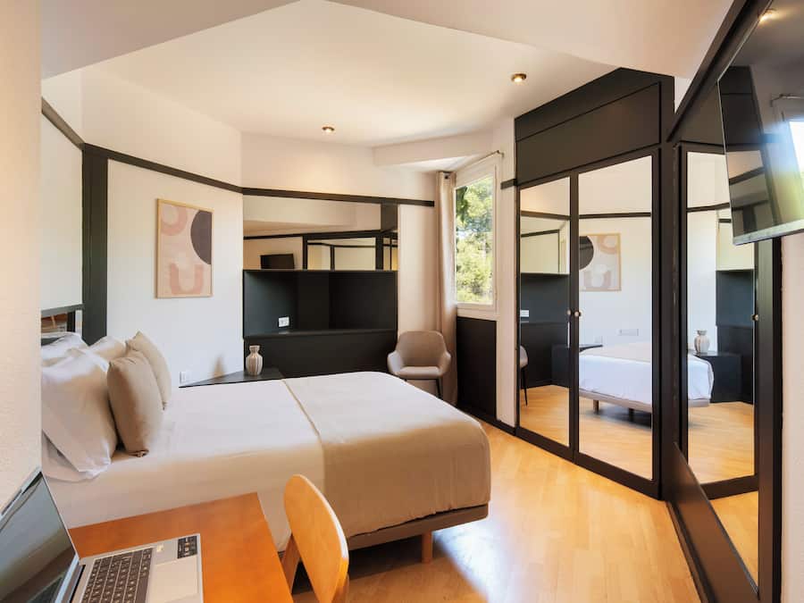 A modern hotel room with a bed, a desk with a laptop, a chair, and a large mirror.