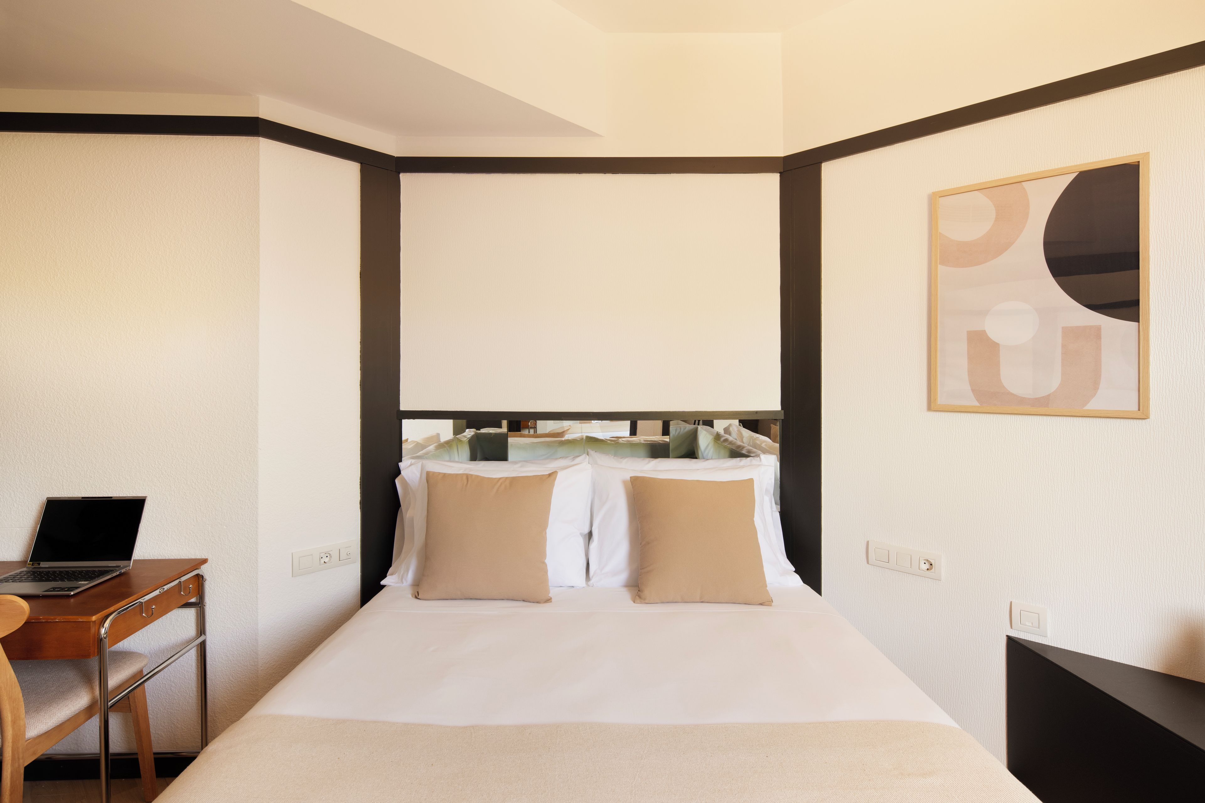 A hotel room with a bed, a desk with a laptop, and a painting on the wall.