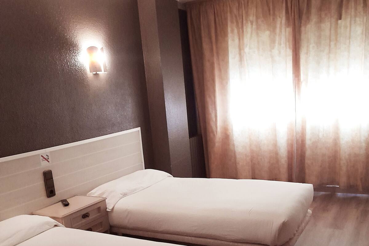 Hotellihuone, jossa on kaksi erillistä sänkyä, yöpöytä, jossa on puhelin, seinään kiinnitetty valaisin ja ikkuna, jossa on verhot.
