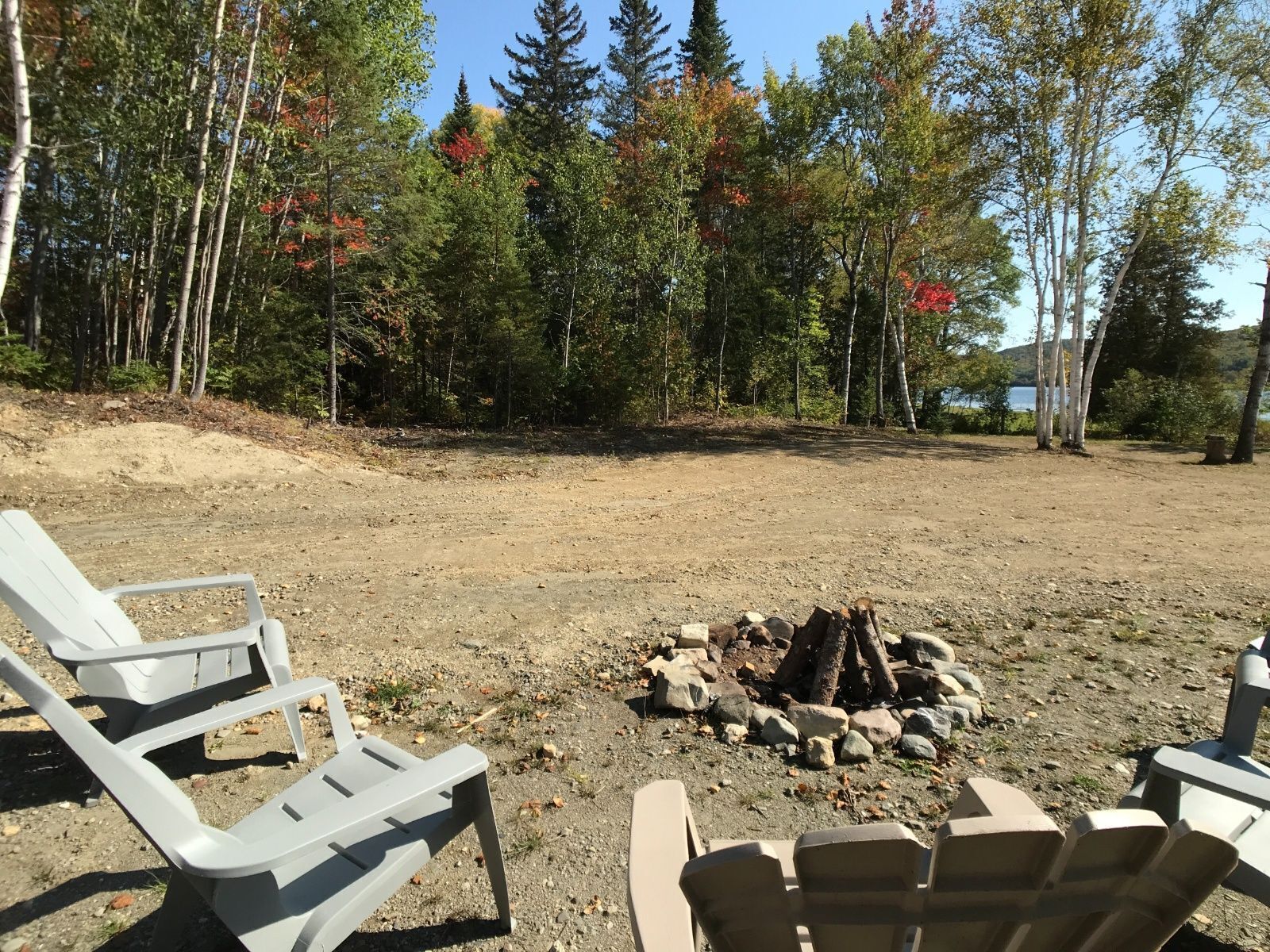 A campsite with two white plastic chairs, a fire pit with logs, and a forested backdrop.