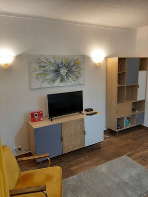 A living room with a flat-screen TV on a wooden cabinet, a yellow armchair, a grey rug, and a bookshelf with multiple compartments.