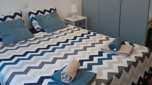 Uma cama bem arrumada com colcha estampada, uma mesa de cabeceira com abajur e um guarda-roupa em um quarto com paredes lisas.