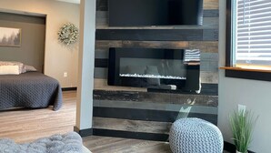 Ein modernes Schlafzimmer mit einem großen Flachbildfernseher an einer Holzwand, einem Bett mit grauer Tagesdecke und einem kleinen runden Hocker.