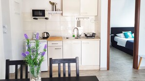 A compact kitchen with white cabinets, a microwave, and a sink. There's a dining table with chairs and a vase of flowers. A bed with a blue and white pillow is visible through an open doorway. - Spectacular View only 5 min. walk from beach, restaurants, attractions and more! (Korčula)