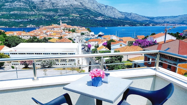 A balcony with two chairs and a table, overlooking a coastal town with buildings and a mountain in the background. - Spectacular View only 5 min. walk from beach, restaurants, attractions and more! (Korčula)