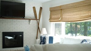 Un salon avec un mur en briques blanches, un téléviseur à écran plat fixé au-dessus d’une cheminée, un canapé blanc agrémenté de coussins à motifs, une table basse en bois et une fenêtre avec un store en bambou.