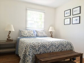 A bedroom with a bed, two bedside lamps, a window, and four framed pictures on the wall. - Summer in the Hamptons (Westhampton Beach)