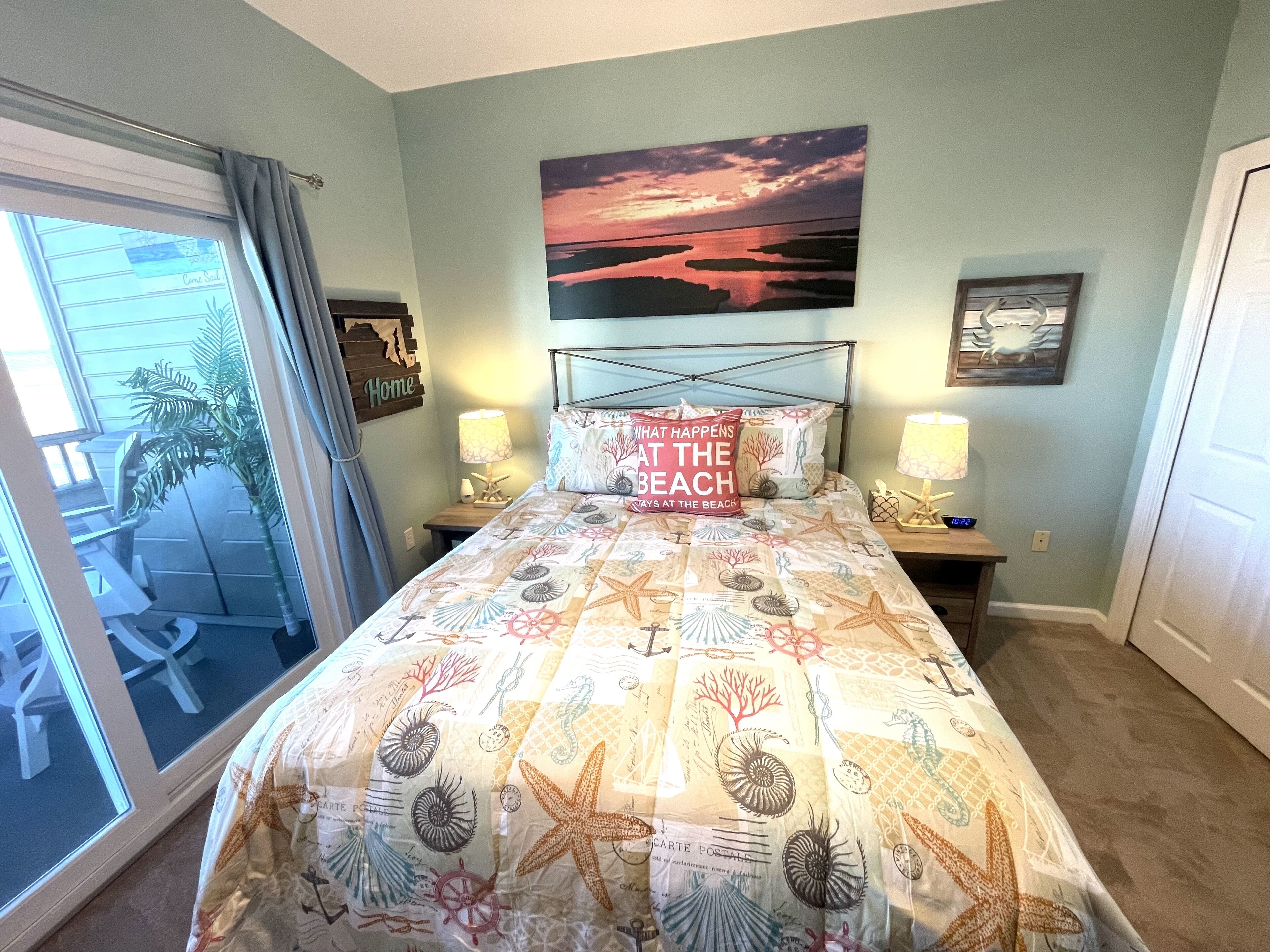 Ein Schlafzimmer mit einem Bett, zwei Nachttischlampen, einem Wandbild und Blick durch eine Schiebetür in einen Außenbereich.