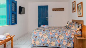 Un dormitorio con cama, mesita de noche de madera, escritorio, silla, ventana con contraventanas azules, puerta azul y televisor montado en la pared.