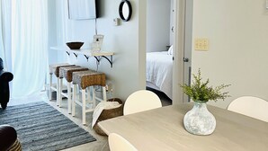 Una habitación de hotel moderna con zona de comedor que incluye una mesa de madera y sillas blancas, un mueble blanco peque ño y un taburete de mimbre.
