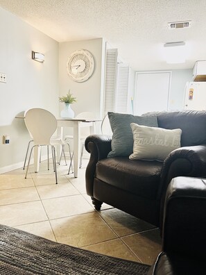 Uma sala de estar com um sofá de couro preto, uma mesa de jantar branca e uma cadeira branca. Há um relógio na parede e uma pequena planta sobre a mesa.