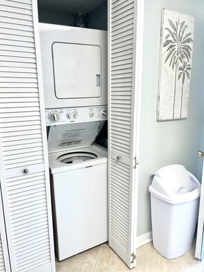 A compact laundry room with a front-loading washer and dryer, a trash bin, and a door with a louvered design.