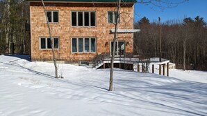 A two-story wooden cabin with a green roof, a covered deck, and a snow-covered ground. - Large relaxing house in 25 acres of private woods with lake access. (Spencer)