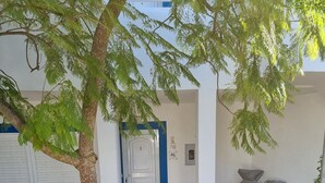 Un jardín ajardinado con diversas plantas y rocas, una puerta blanca y una zona de estar cubierta con un toldo azul.