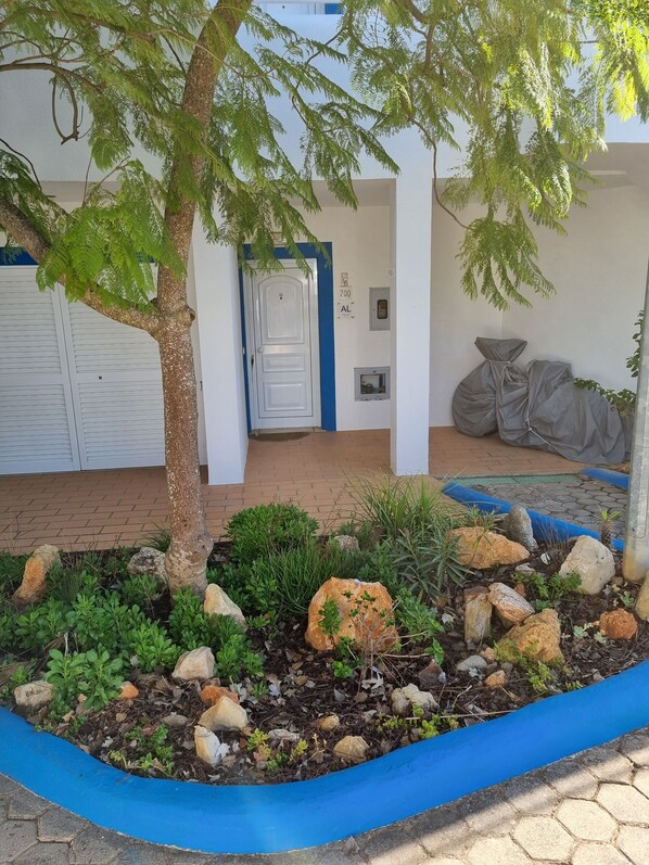 A landscaped garden area with a variety of plants and rocks, a white door, and a covered seating area with a blue awning.