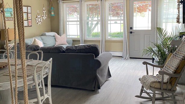 Une pièce chaleureuse avec un plafond en bois, un canapé gris, un fauteuil à bascule et une table à manger entourée de chaises en corde.