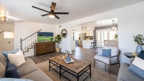 A spacious living room with a wooden coffee table, a sofa, and a TV. There's a ceiling fan, a staircase, and a kitchen area visible in the background. - Old Town Scottsdale | Heated Pool & Putting Green (Scottsdale)