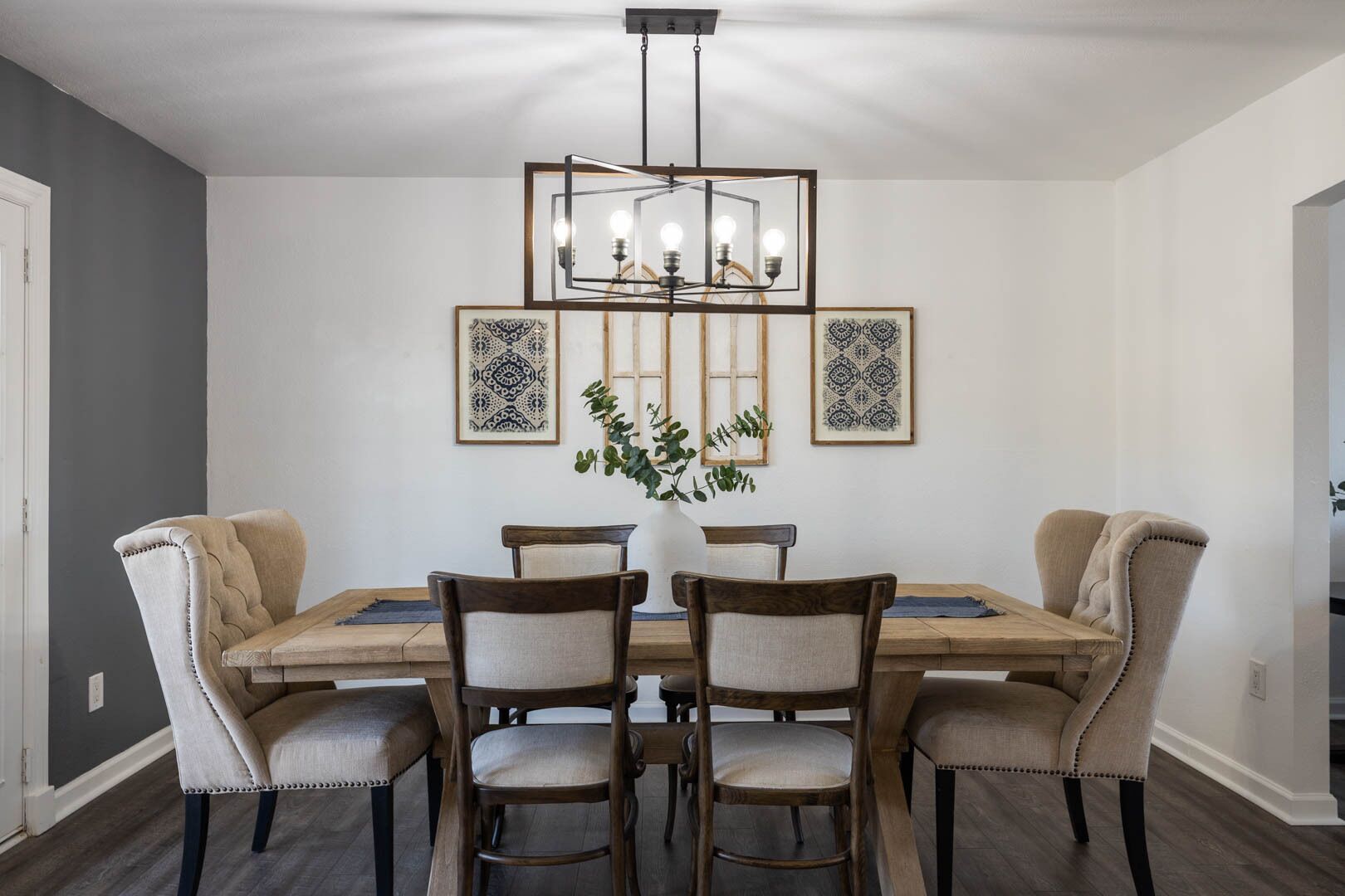 A dining room with a wooden table, four upholstered chairs, a chandelier, and two framed wall art pieces.