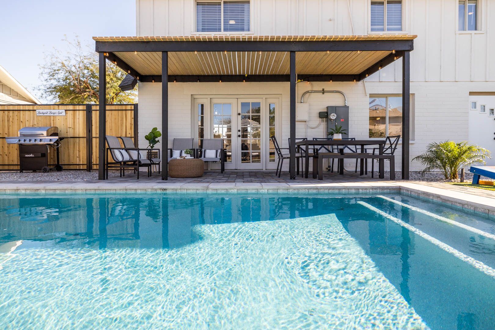 A pool area with a covered patio, outdoor furniture, and a grill.