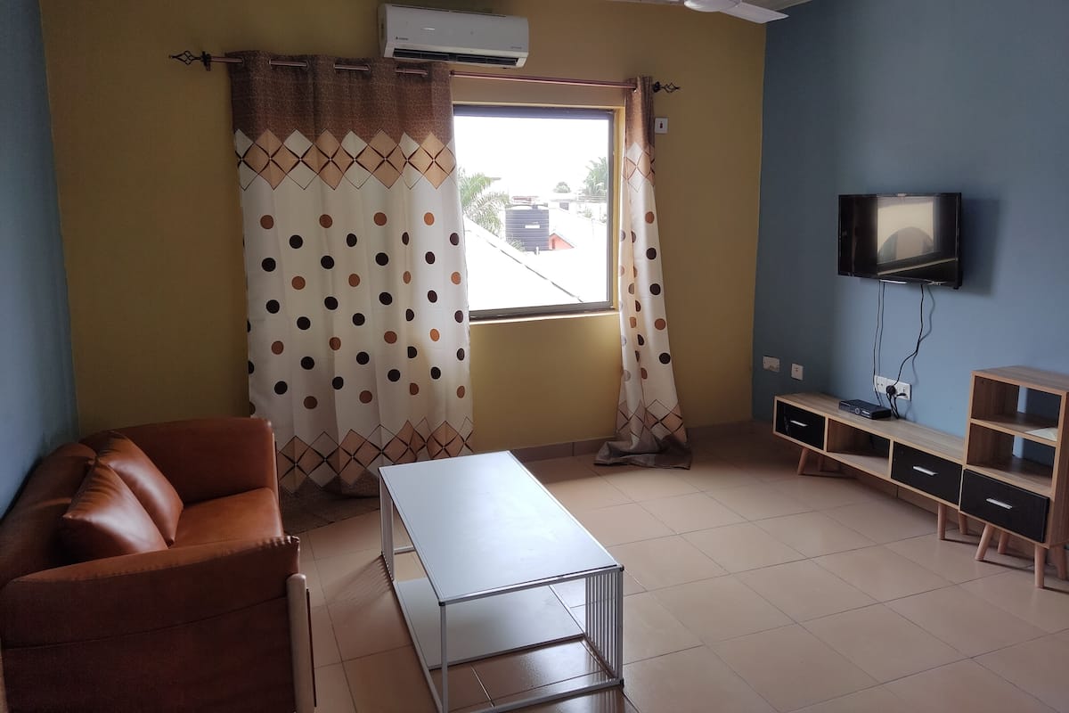 A living room with a brown sofa, a white table, a TV on the wall, and a window with curtains.