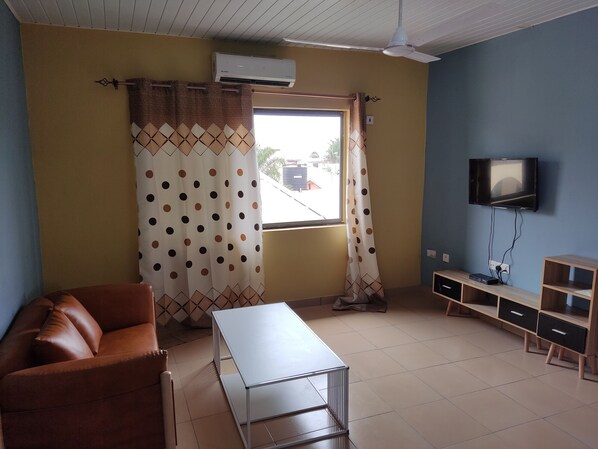 Uma sala de estar com um sofá marrom, uma mesa branca, uma televisão na parede e uma janela com cortinas.