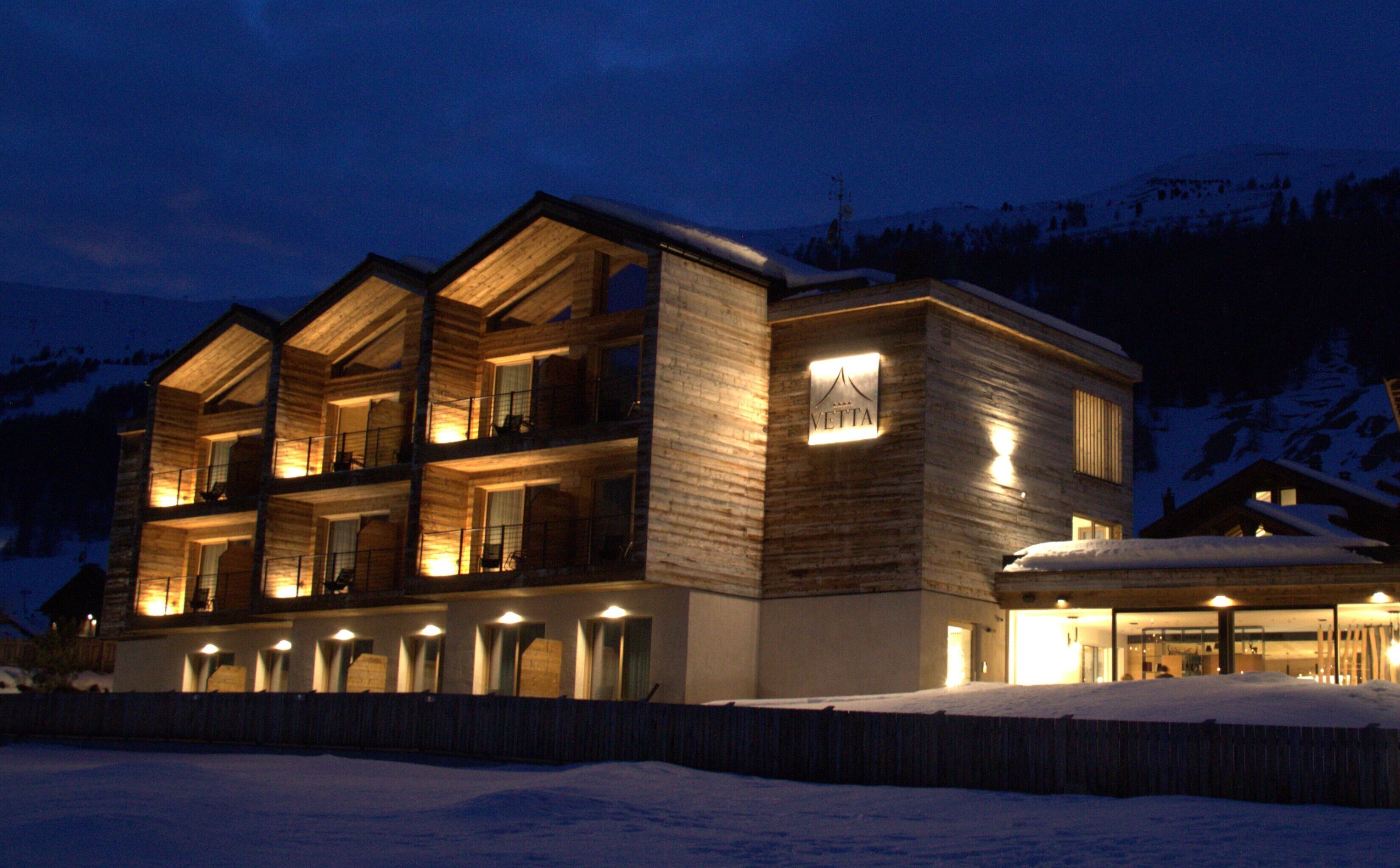 Un edificio de madera de varios pisos, con balcones, ventanas iluminadas y un letrero que dice "VETTA".