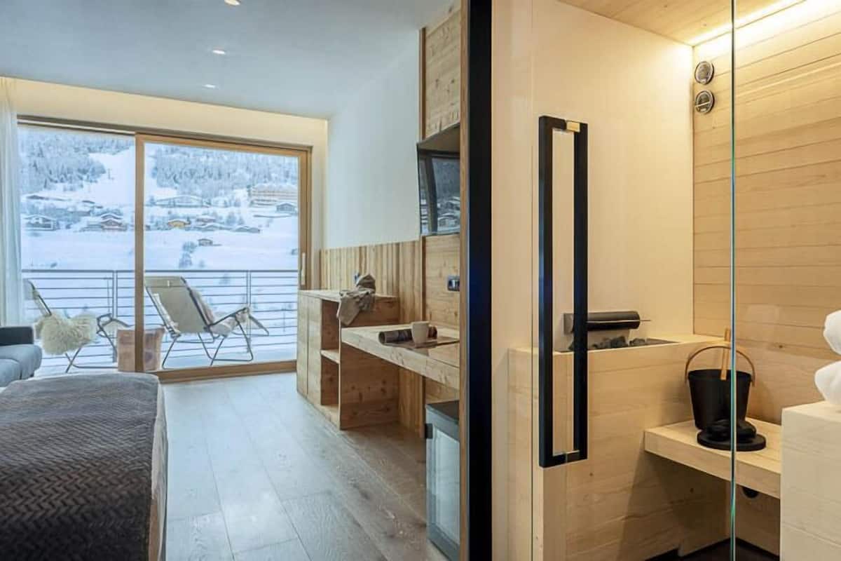 Una habitación de hotel moderna con un amplio ventanal corredera que da a un balcón, un escritorio de madera, una cama con manta gris y un armario de madera.