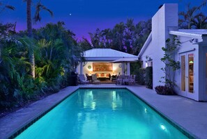 Una piscina con zona de patio, rodeada de exuberante vegetación y una casa moderna con fachada blanca.