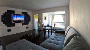 Un salon comprenant un grand canapé d’angle, un téléviseur à écran plat fixé au mur, un coin repas avec une table et des chaises, et une fenêtre avec des rideaux.