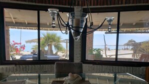 Un comedor con una mesa de vidrio, una estatua y vista a una playa con palmeras y una cabaña de techo de paja.