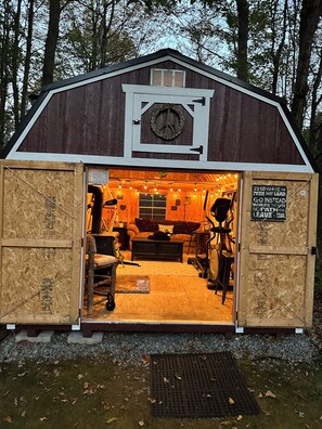 Una rustica rimessa in legno con un orologio sulla facciata, un interno accogliente con un divano e un tavolino, e un cartello motivazionale sulla porta.
