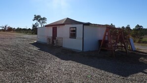 A small, single-story building with a red door and a window, surrounded by gravel and trees. - Cozy casita: Bastrop state park view (Bastrop)
