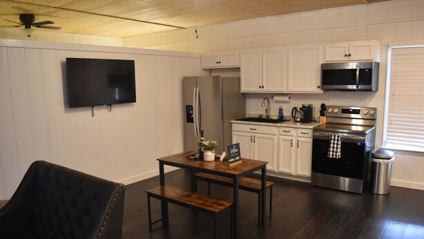 A modern kitchen with white cabinets, a stainless steel refrigerator, a microwave, and an oven. There's a wooden dining table with benches, a black leather chair, and a TV mounted on the wall. - Lovely one room barndominium - The Bastrop Barndo  (Bastrop)