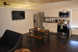 A modern kitchen with white cabinets, a stainless steel refrigerator, a microwave, and an oven. There's a wooden dining table with benches, a black leather chair, and a TV mounted on the wall.