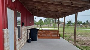 A covered outdoor area with a concrete floor, a wooden bench, a trash bin, and a red building with a white door. - Lovely one room barndominium - The Bastrop Barndo  (Bastrop)
