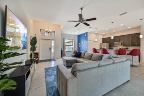 A modern living room with a sectional sofa, a dining area with red chairs, and a kitchen with dark cabinets and a microwave.