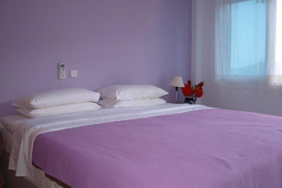 A bed with a purple bedspread, white pillows, and a nightstand with a lamp and flowers.