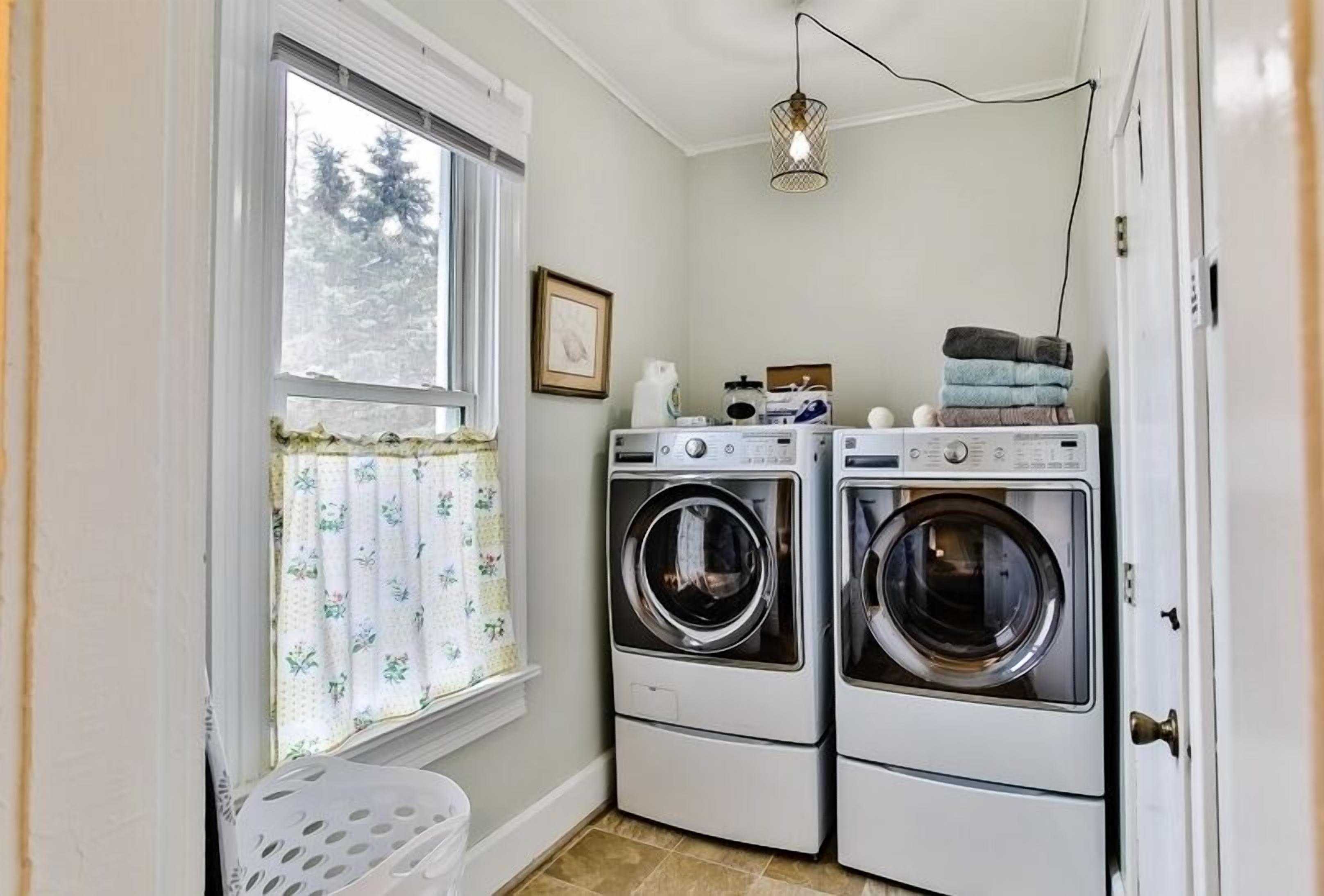 A laundry room with two front-loading washing machines, a drying rack, and a window with floral curtains.