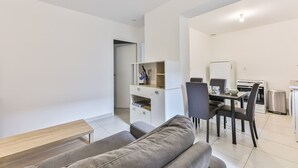 Un salon moderne et bien éclairé, avec un canapé gris, une table basse en bois et un coin repas comprenant une table blanche, des chaises noires et un espace cuisine équipé d’appareils électroménagers.