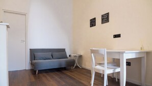 Ein Zimmer mit grauem Sofa, einem weißen Tisch mit Stuhl und einem kleinen Nachttisch mit Lampe.