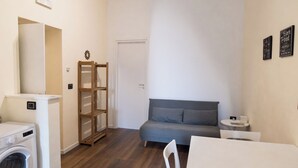 Ein kompakter Wohnraum mit Waschmaschine, einem grauen Sofa, einem kleinen Tisch und einer Holzhängeregal.