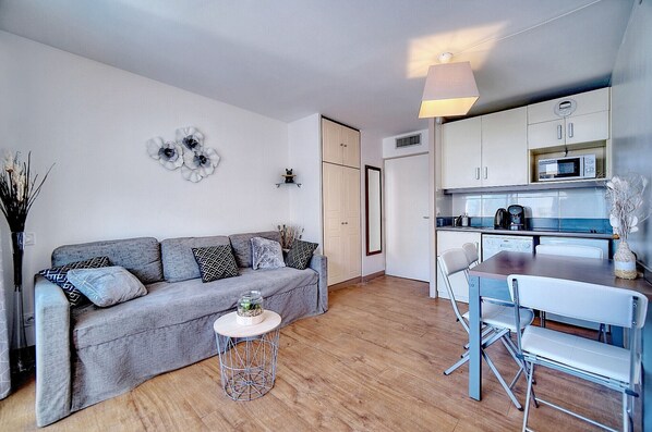 A compact living space with a grey sofa, a small round table, a dining table with chairs, a kitchen area with cabinets and a microwave, and a large mirror.