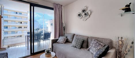 A living room with a grey sofa, patterned cushions, a round side table, and a view of a building through a large window.