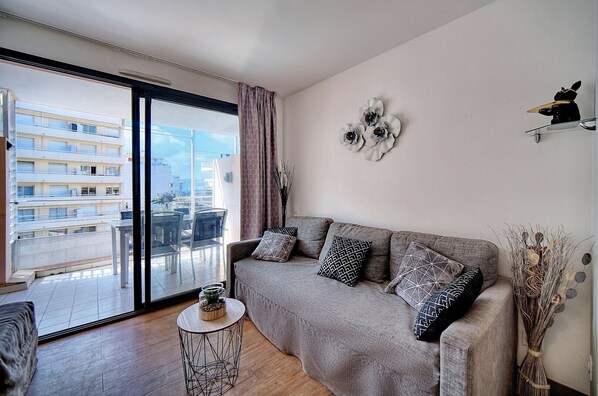 A living room with a grey sofa, patterned cushions, a round side table, and a view of a building through a large window.